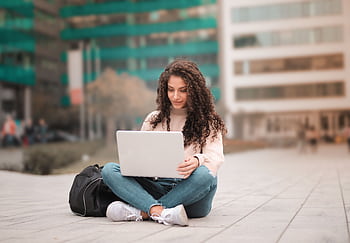 A woman with curly hair using a laptop while sitting outdoors, showcasing modern remote work lifestyle. HD wallpaper