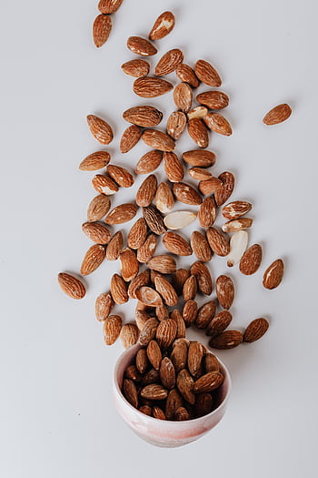 Top view of scattered raw almonds around a pink ceramic bowl on a clean white surface. HD phone wallpaper
