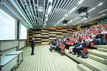 A speaker delivers a presentation to an attentive audience in a modern auditorium setting. HD wallpaper