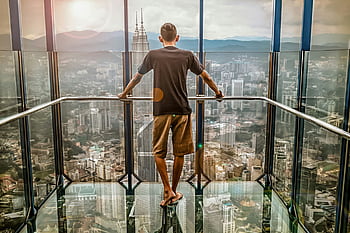 A man stands barefoot on a glass deck high above Kuala Lumpur, enjoying the stunning cityscape view. HD wallpaper