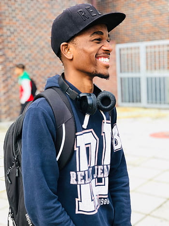 Smiling young man wearing cap and heads, standing with backpack outdoors. HD phone wallpaper
