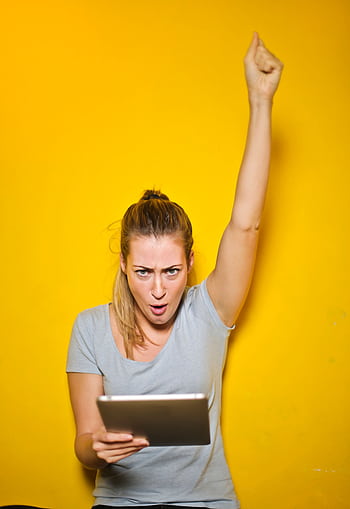 Joyful woman raises arm in celebration while holding a tablet against a vibrant yellow backdrop. HD phone wallpaper