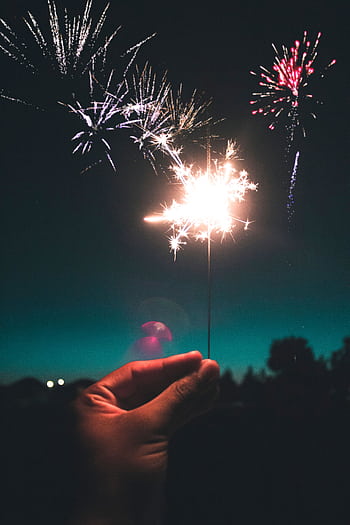Close-up of a hand holding a sparkler with fireworks illuminating the night sky. HD phone wallpaper