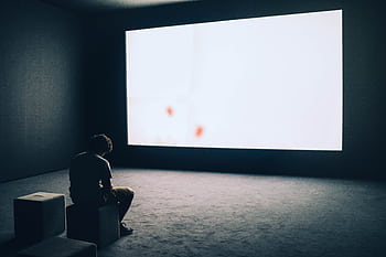 A man sits alone in a dark room with a large white screen, depicting solitude and contemplation. HD wallpaper