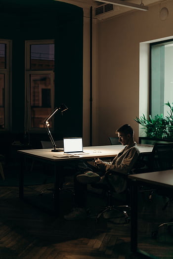 A young man using a laptop at night in a dimly lit office space, focused on his work. HD phone wallpaper