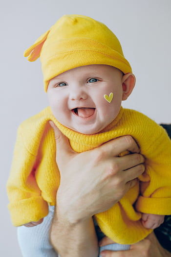 Adorable baby wearing a yellow beanie with a heart sticker, smiling joyfully indoors. HD phone wallpaper
