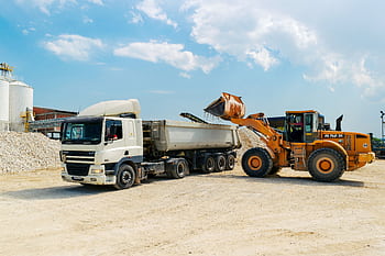 Excavator loading materials into a heavy-duty truck at a sunny construction site. HD wallpaper