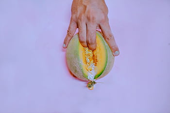 A hand gently touches a ripe melon on a pastel pink backdrop, creating a vivid and conceptual . HD wallpaper