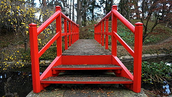 A vibrant red wooden bridge crossing a serene stream in a tranquil autumn park. HD wallpaper