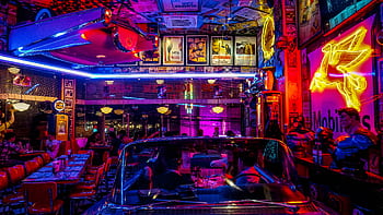 A lively retro diner bustling with people, neon lights, and classic car decor, set at night. HD wallpaper