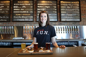 Smiling bartender in a bar serving craft beer flights with taps and menu board in the background. HD wallpaper