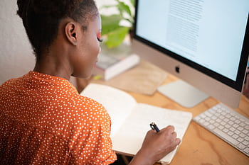 A woman in a polka-dot shirt writes in a notebook at her desk with a computer screen visible. HD wallpaper