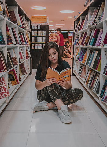 Young woman sitting on the floor and reading a book in a modern bookstore with shelves full of colorful books. HD phone wallpaper