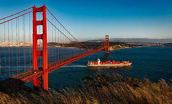 Golden Gate Bridge arches over San Francisco Bay as a cargo ship passes beneath during sunset. HD wallpaper