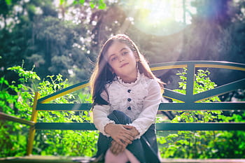A young girl in stylish attire sitting on a bench in a sunny outdoor garden setting. HD wallpaper