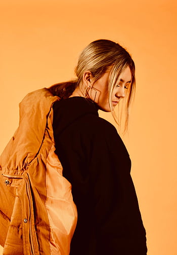 Portrait of a woman in a yellow jacket, posing thoughtfully against a warm orange backdrop. HD phone wallpaper