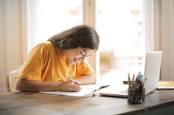 Young woman studying with a laptop and notepad at home, smiling and focused. HD wallpaper