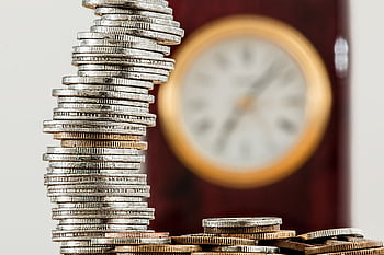 A close-up of stacked coins with a blurred clock, symbolizing time and money relationship. HD wallpaper