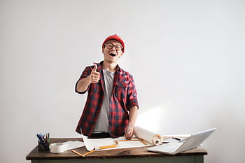 Cheerful male architect in a red hardhat and plaid shirt giving thumbs up at his desk. HD wallpaper