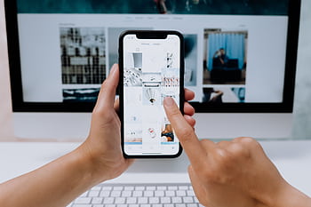 Hands holding a smart while browsing a social media app in front of a computer. HD wallpaper