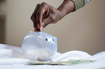 Close-up of a person's hand placing coins into a transparent piggy bank to save money. HD wallpaper