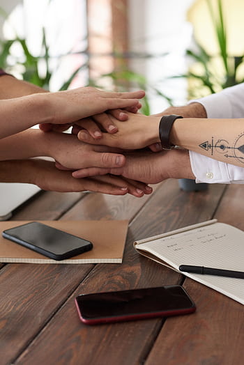 Hands from a diverse team stack on a table symbolizing unity and teamwork in a modern office setting. HD phone wallpaper