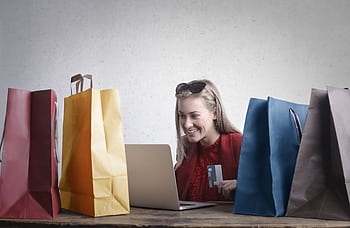 A joyful woman shopping online using a laptop with colorful shopping bags surrounding her. HD wallpaper
