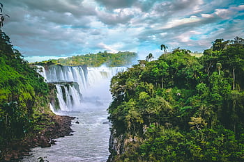 Breathtaking view of towering waterfalls surrounded by vibrant green jungle and dramatic clouds. HD wallpaper