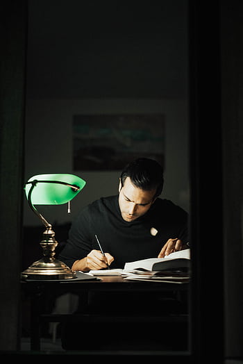 Focused man writing at desk under a green lamp, immersed in study. HD phone wallpaper