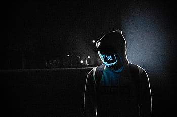 A mysterious figure wearing a LED mask and hoodie stands illuminated in a dark, foggy street at night. HD wallpaper