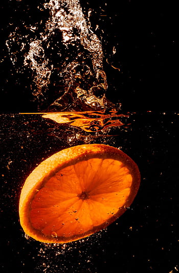 Orange slice creates a splash in water on black background, capturing freshness and energy. HD phone wallpaper