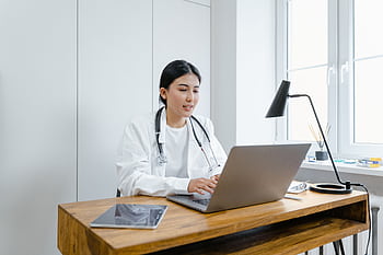 A female doctor in a white coat uses a laptop for an online consultation from her office. HD wallpaper