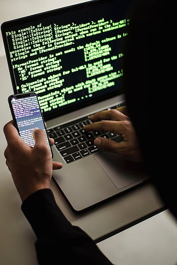 A hooded figure engaged in hacking using a laptop and smart in low light. HD phone wallpaper