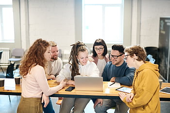 A diverse team of professionals collaborates happily around a laptop in a bright indoor setting. HD wallpaper