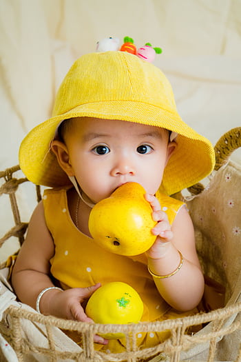 Cute baby in a yellow hat holding a pear, sitting in a basket with a playful expression. HD phone wallpaper