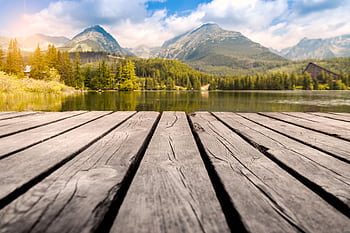 Calm mountain lake with a wooden dock, surrounded by lush forests and mountains under a clear sky. HD wallpaper