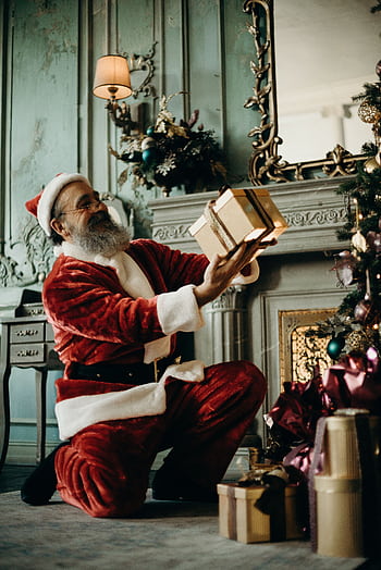 Santa Claus joyfully holding gifts by a decorated Christmas tree indoors. HD phone wallpaper