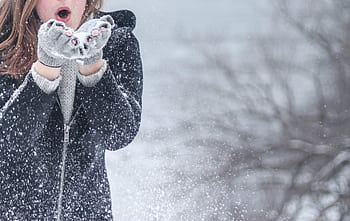 Woman in cozy winter clothing blowing snowflakes with excitement outdoors in a snowy setting. HD wallpaper