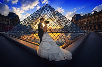 Couple embraces before the Louvre Pyramid during a stunning sunset in Paris. HD wallpaper