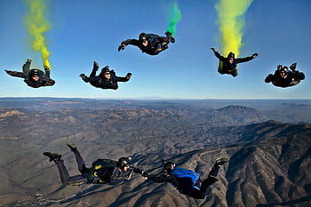 Dynamic aerial shot of skydivers performing stunts with colorful smoke trails over mountainous terrain. HD wallpaper