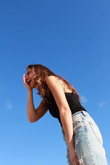A young woman in casual attire laughing outdoors against a vibrant blue sky, radiating happiness. HD phone wallpaper
