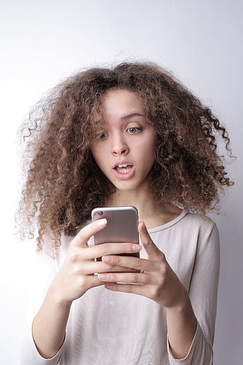 A young woman with curly hair looks surprised while reading something on her smart indoors. HD phone wallpaper