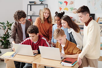 A group of diverse students collaborating on laptops in a classroom setting, guided by a teacher. HD wallpaper