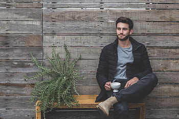 Casual man sitting on a bench with a coffee cup against a wooden wall. HD wallpaper