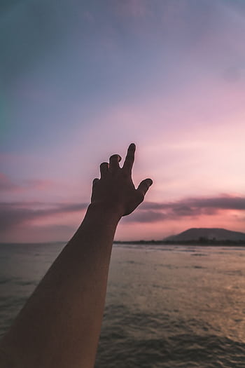 A silhouetted hand stretches upward against a colorful sunset sky over the ocean, evoking a sense of wonder and exploration. HD phone wallpaper