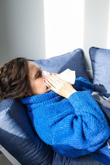 A woman in a blue sweater uses a tissue, lying indoors, suggesting illness or flu symptoms. HD phone wallpaper