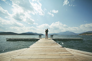 A father embraces his child on a wooden dock by a scenic lake and mountains under a clear sky. HD wallpaper