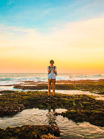 A man stands on mossy rocks by the ocean, praying during a vibrant sunset, creating a serene and introspective scene. HD phone wallpaper
