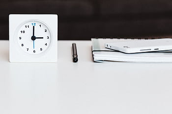 A minimalist office desk setup with a clock, pen, smart, and notebook, conveying time management and organization. HD wallpaper