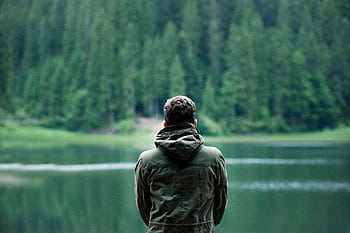 A person in a green jacket stands by a serene forest lake, capturing calming nature. HD wallpaper